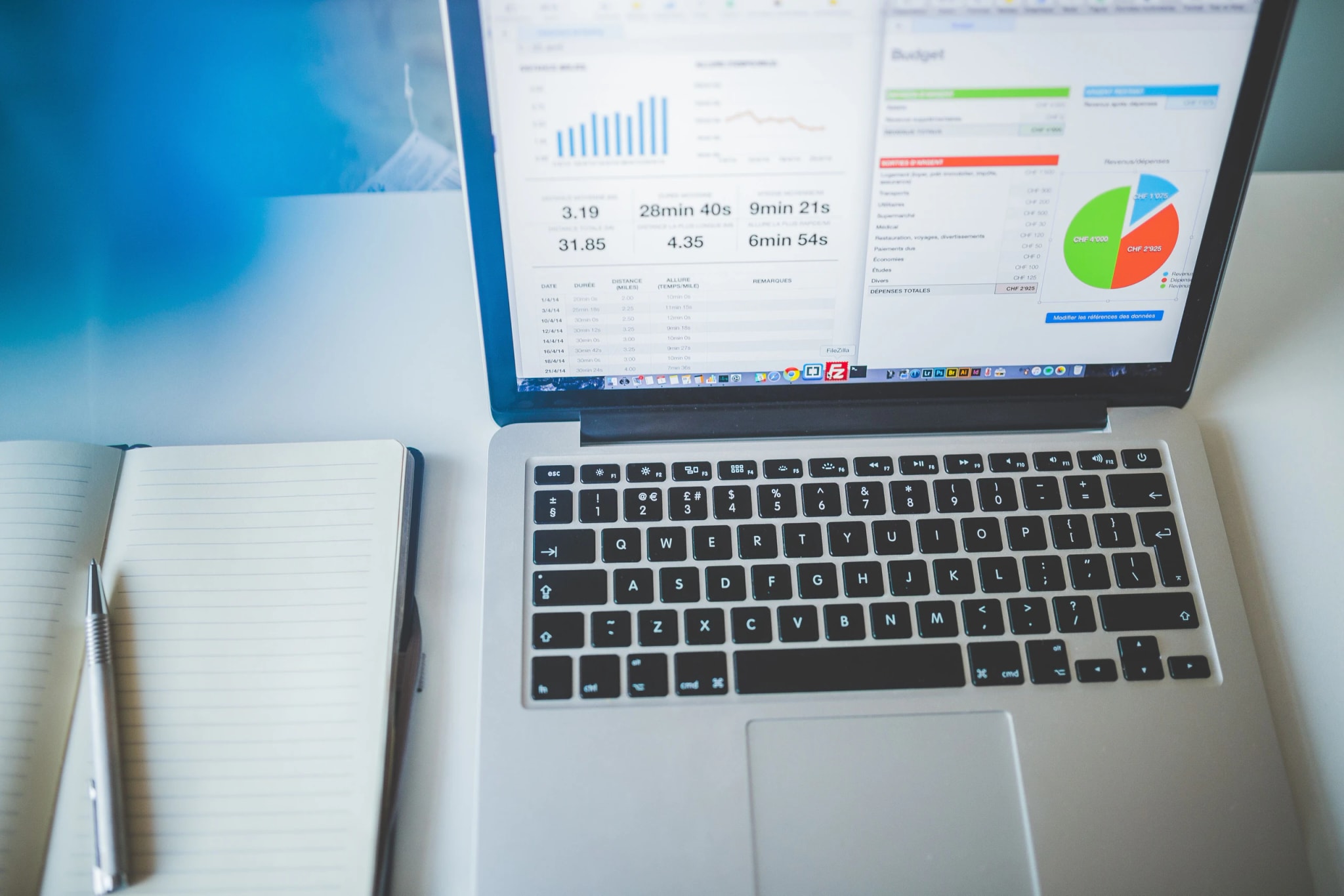 A laptop showing a budget spreadsheet and charts, open beside a ruled paper notebook and a pen.
